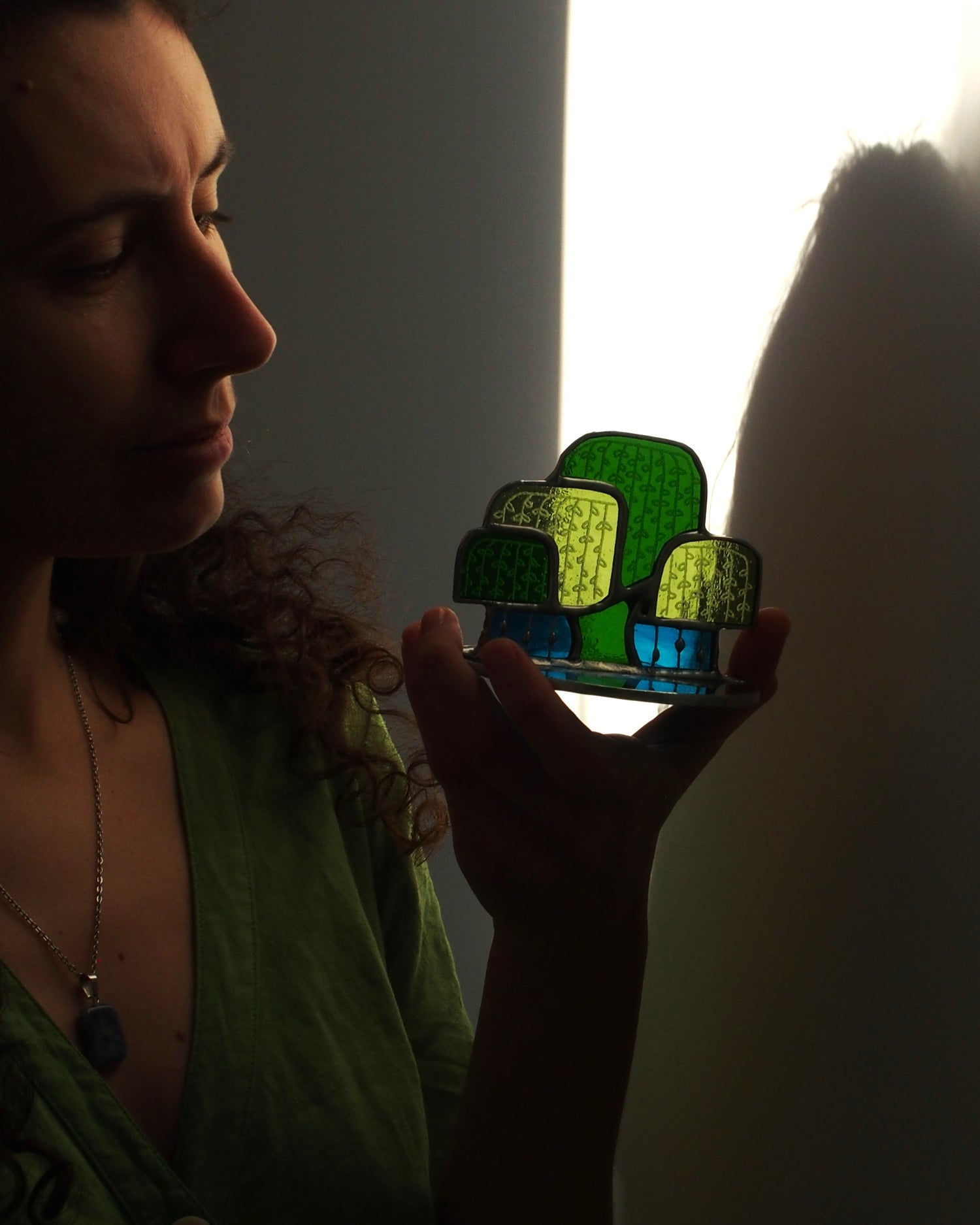 Artisane tenant à la main un vitrail. Celui-ci représente un saule pleureur, dans des tons vert et bleu. Il est éclairé par un rayon de lumière. 